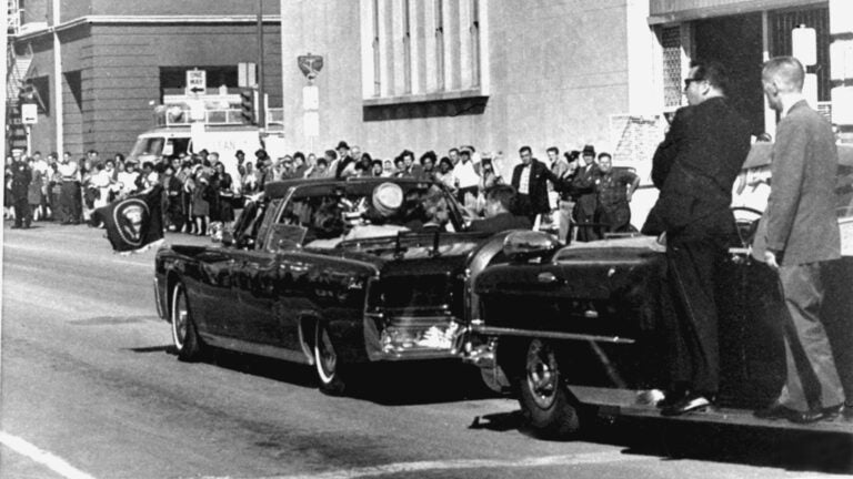 Secret servicemen standing on running boards follow the presidential limousine carrying President John F. Kennedy.