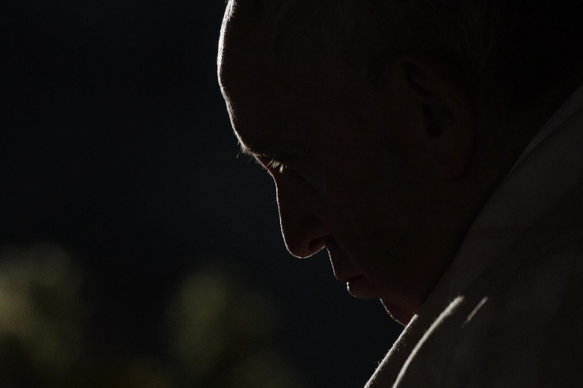 Pope Francis shown in profile bowing his head in the dark.