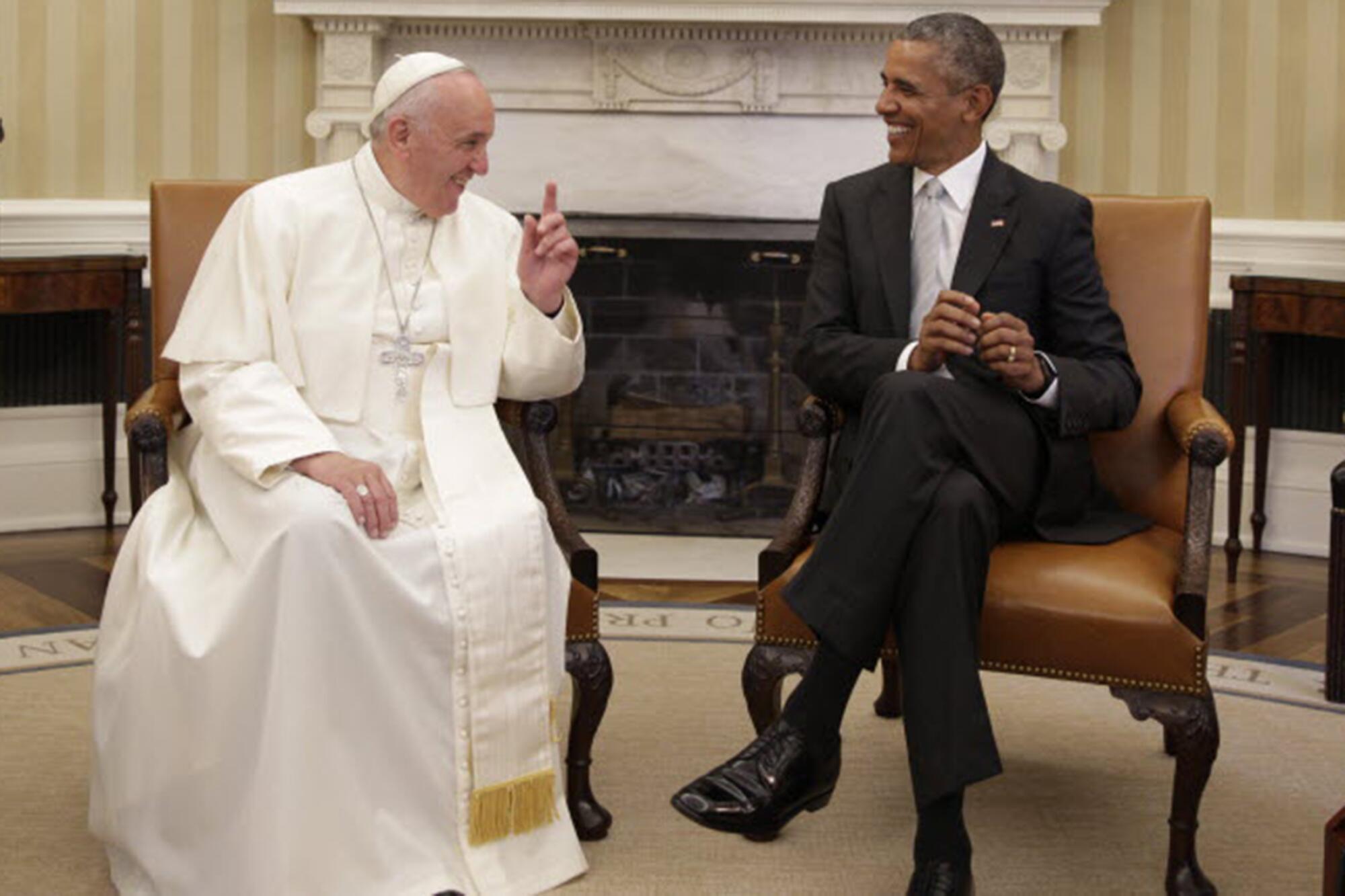 A man in white skullcap and religious vestments smiles as he speaks to another man in a dark suit, also seated