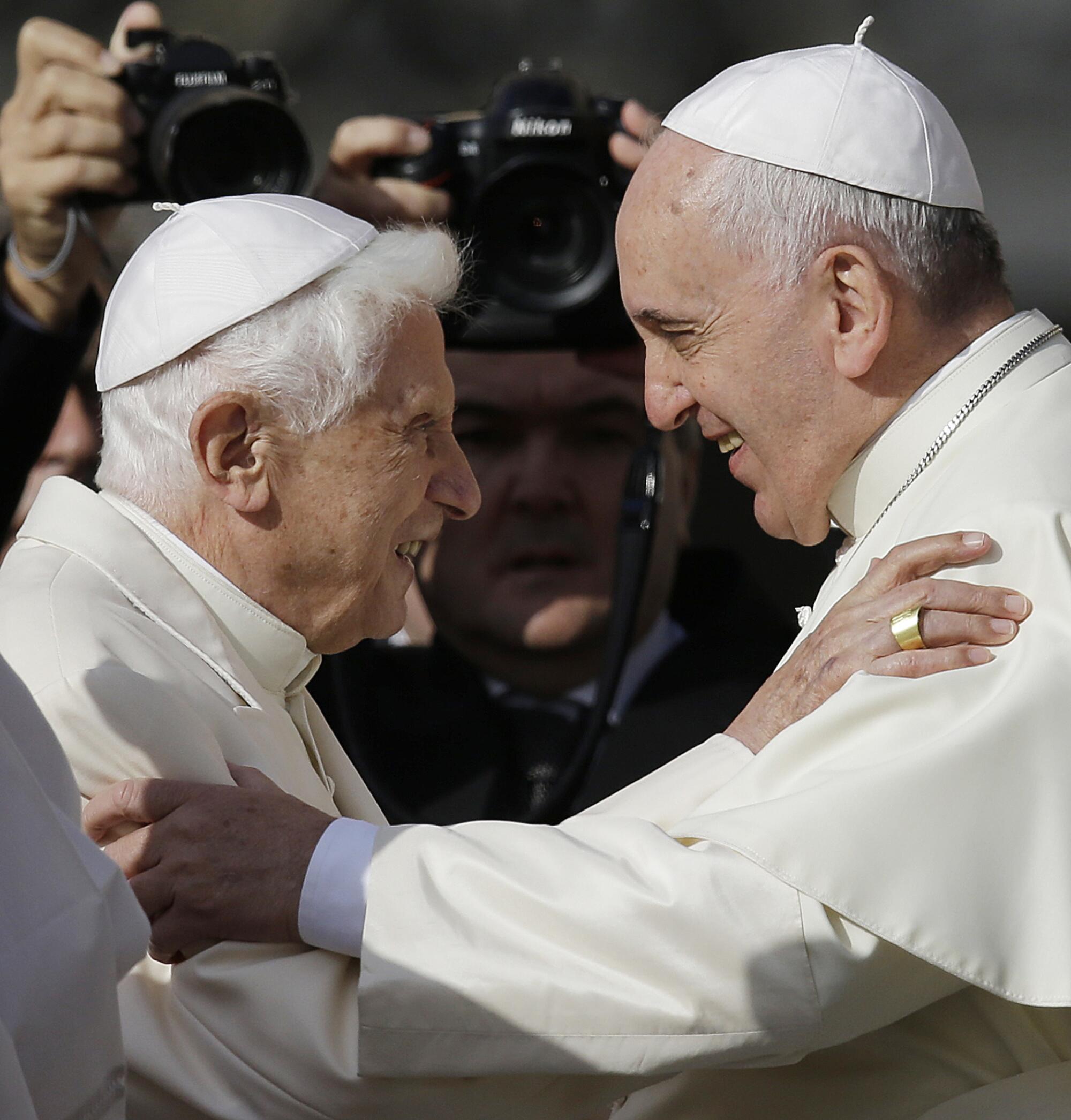 Two smiling men with gray hair, in white skullcaps and robes, embrace.