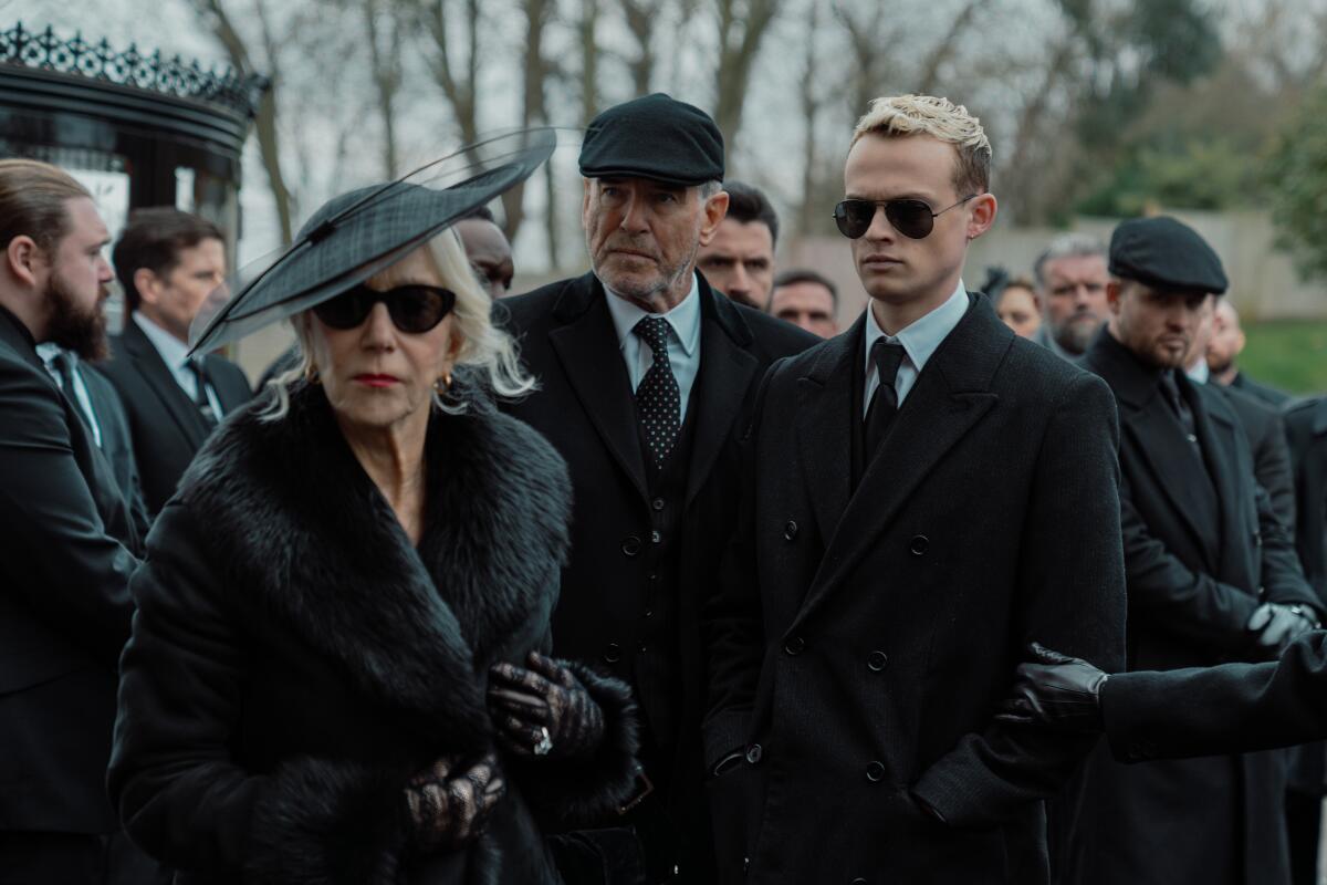 A woman in black fur coat and hat stands next two men in dark suits and coats at a funeral.