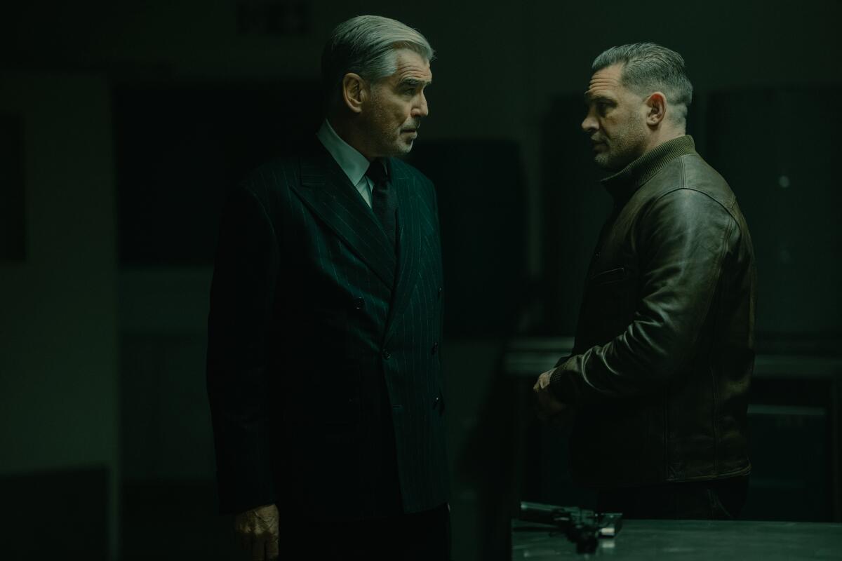 A man in a dark suit looks at a man in a dark leather jacket as they stand near table with a gun on it.