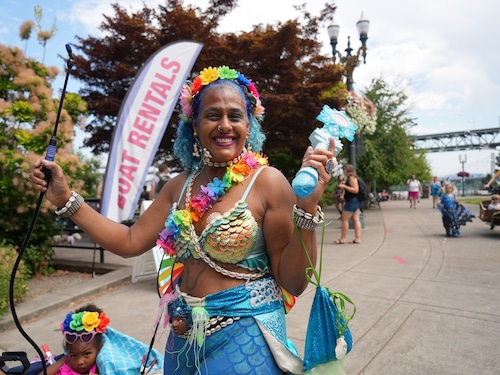 woman dressed like a mermaid with a headband of rainbow colored flowers smiles and holds a bubble making wand