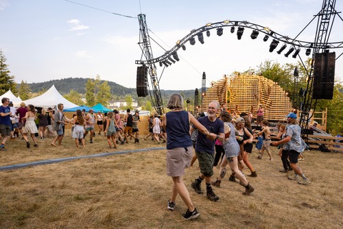People are partaking in a square dance at a music festival. Couples are dosi-do-ing. There is a wooden stage in the background, and they are in a light brown field.