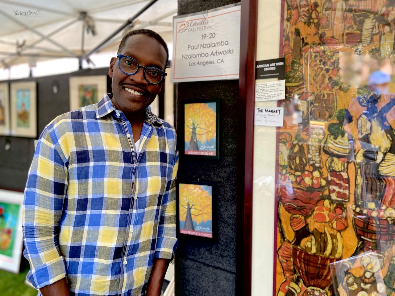 A persoN stands in front of a display of colorful paintings at an outdoor art exhibit. A placard reads ‘Paul Nzalamba, Nzalamba Artworks, Los Angeles, CA’