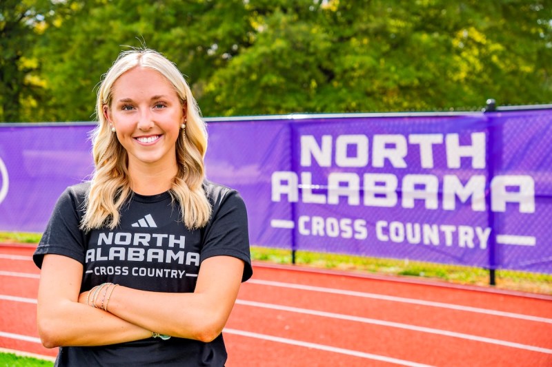 A young woman stands with arms crossed in front of a University of North Alabama Cross Country banner