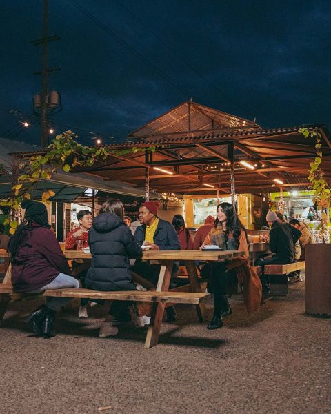 people seated at picnic tables talk and eat with food carts in the background