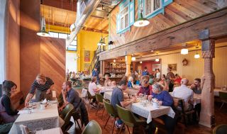 tables full of diners enjoy a meal in a lively, warmly lit restaurant with a bar in the background
