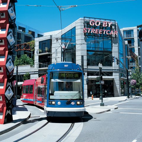 A streetcar passes by a large, red sculpture in front of a building with a neon sign reading, 