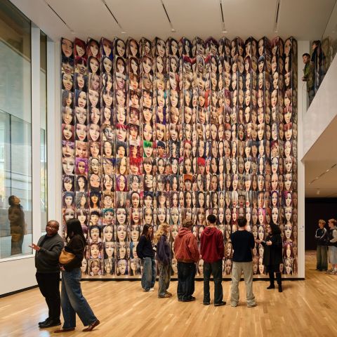 People look at art, including a large, floor-to-ceiling installation featuring many faces, from several different angles: from outside a window, from a ground floor view, and looking over a balcony.