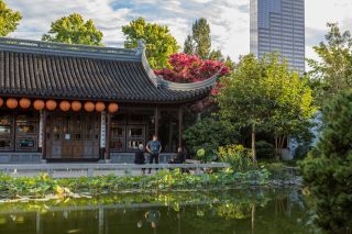 One person stands and two sit between a pond and a building with Chinese architecture.