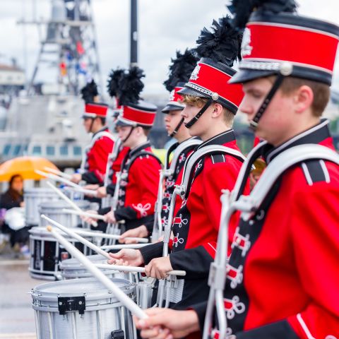 youth drum line of marching band in red costumes and hats walk along bridge in parade