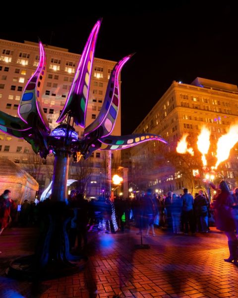 a crowd mingles under a fire display and a lighted purple sculpture