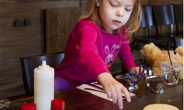 A young girl adds chocolate chips and blueberries to a pancake.