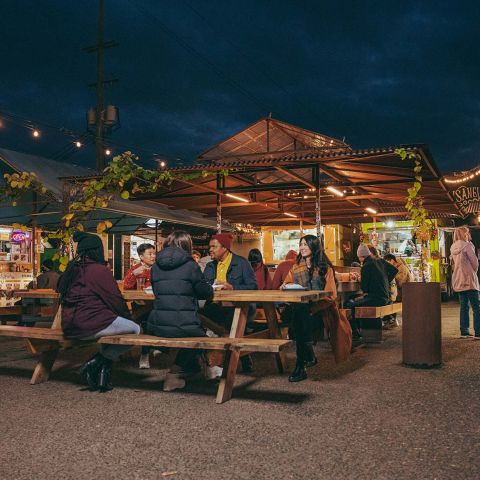 people seated at picnic tables talk and eat with food carts in the background