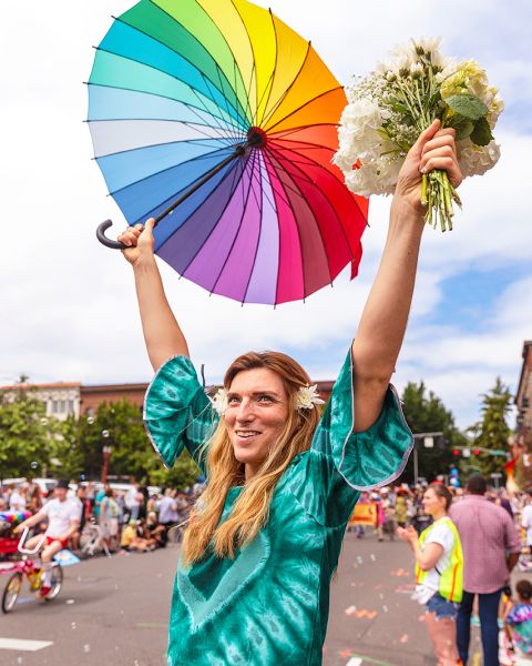 a smiling person at a parade holds a rainbow umbrella and a bouquet of flowers above their head