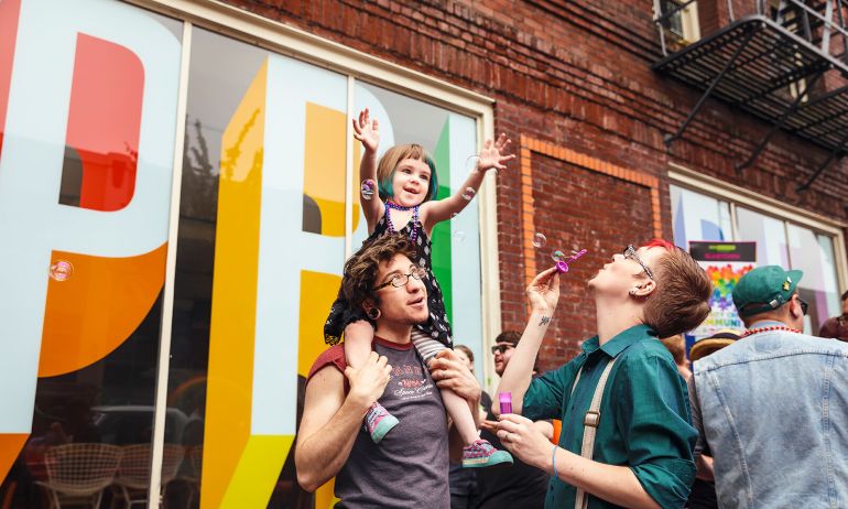 a parent carries their child on their shoulders while the other parent blows bubbles toward them