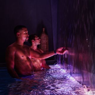 Two men soak in a dark pool lit with purple lights and flanked by a cascading water feature.