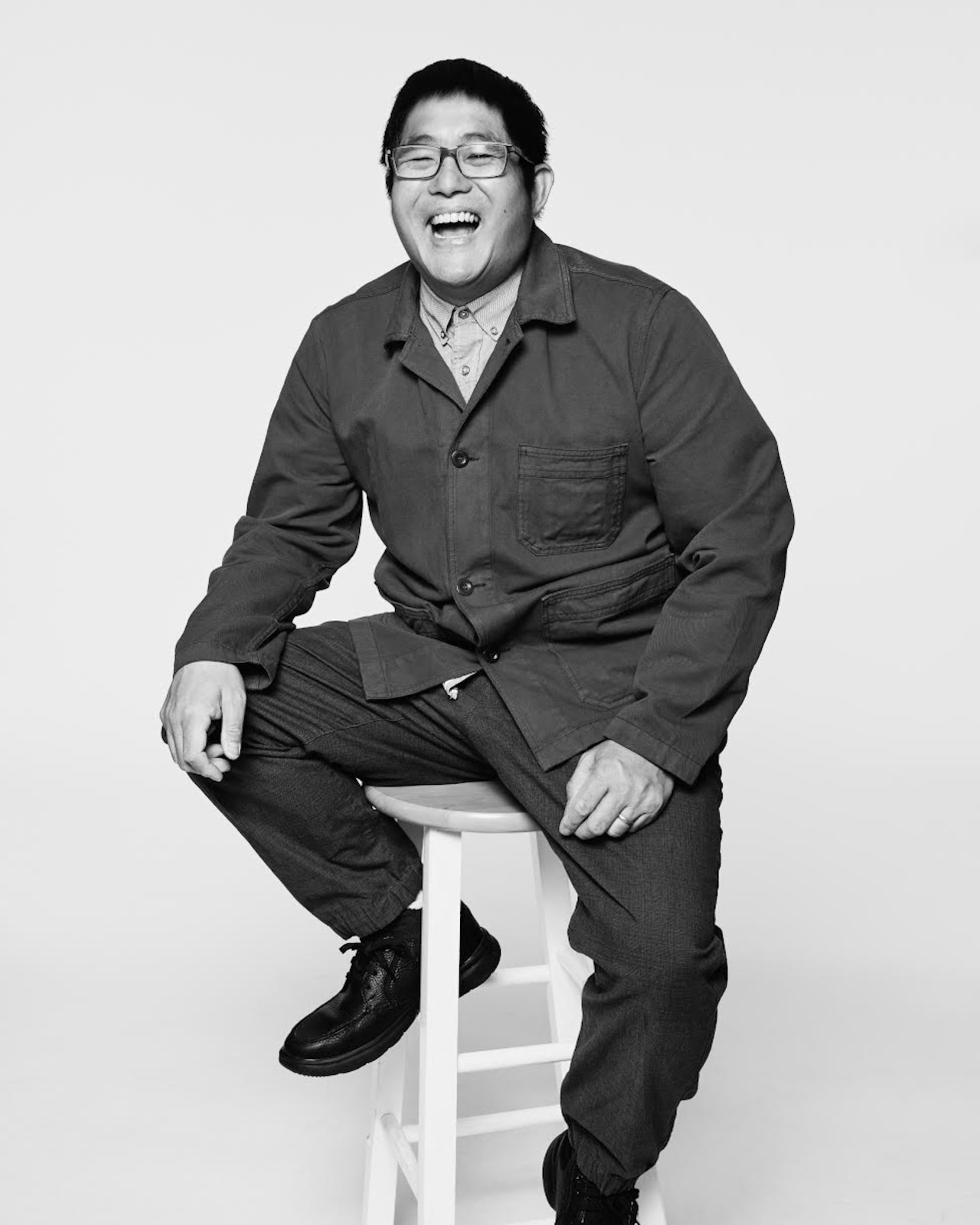 A black and white photograph of Mike Lew sitting on a stool, laughing