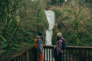 Two people stand at a fence looking toward a waterfall surrounded by wilderness.