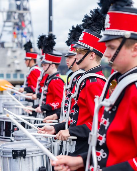 youth drum line of marching band in red costumes and hats walk along bridge in parade