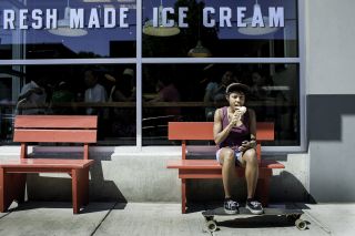 person eats ice cream on red bench with feet on a skateboard in front of storefront of busy ice cream shop