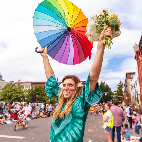 a smiling person at a parade holds a rainbow umbrella and a bouquet of flowers above their head