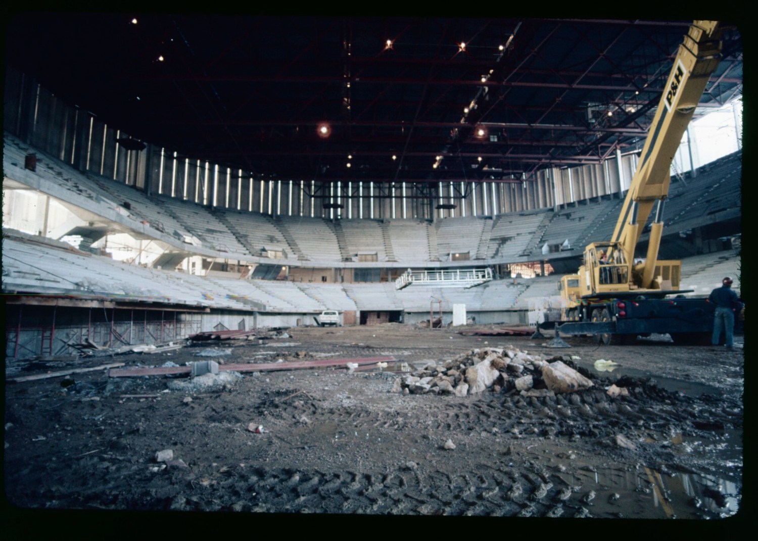 Interior of a large arena under construction or renovation, with exposed seating, debris on the floor, and a yellow P&H crane on the right