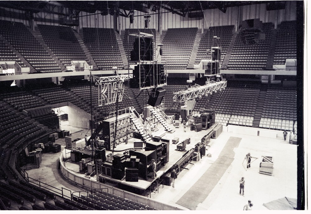 KISS concert stage being assembled in an empty arena, with large speaker towers, rigging, and crew working on the floor
