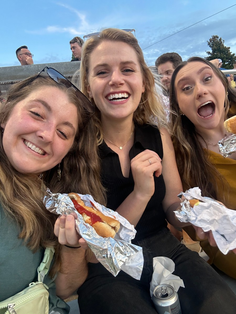 Three friends smiling and holding foil-wrapped hot dogs at an outdoor summer event