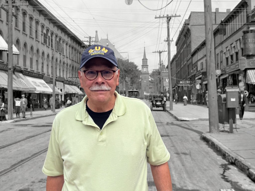 A man with a mustache wearing a yellow t-shirt and ball cap stands in front of a black and white photo of Church Street