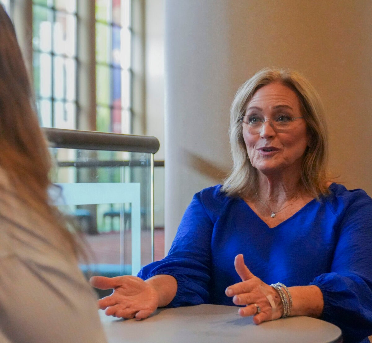 Woman in blue shirt wearing glasses talking to another woman at a table