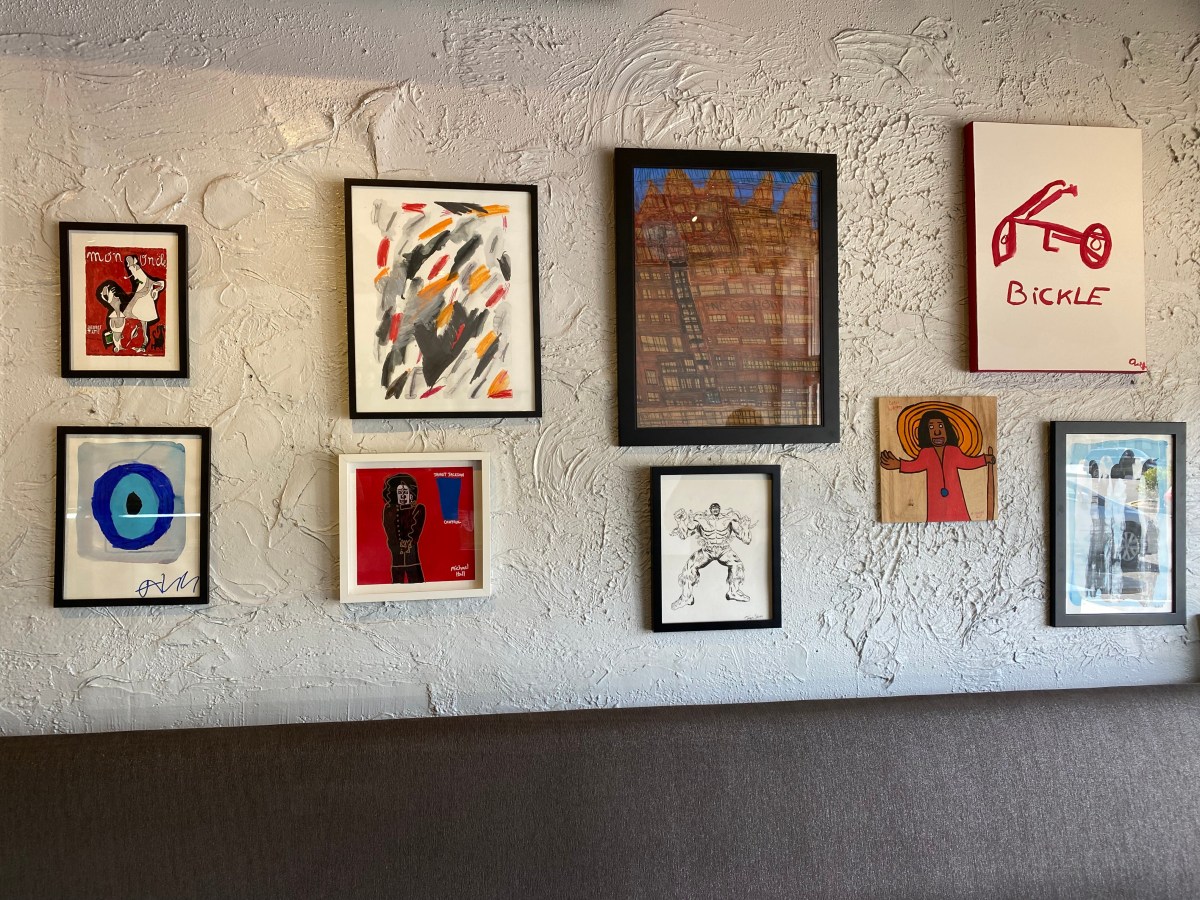A gallery wall displaying diverse Studio By the Tracks artwork, including paintings of a cityscape, abstract brushstrokes, and figurative pieces