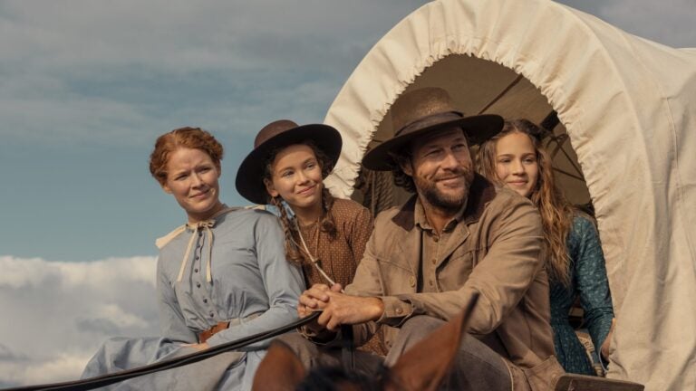 "A man and three women in pioneer-era clothing ride a horse-drawn covered wagon outdoors, under a blue sky with clouds, suggesting a journey or travel in a historical rural setting."