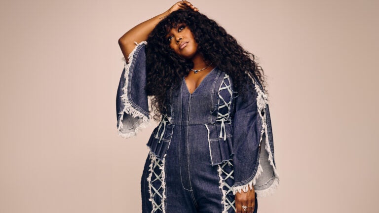 A woman poses in a fringed denim jumpsuit with her hand resting on her head against a plain backdrop.