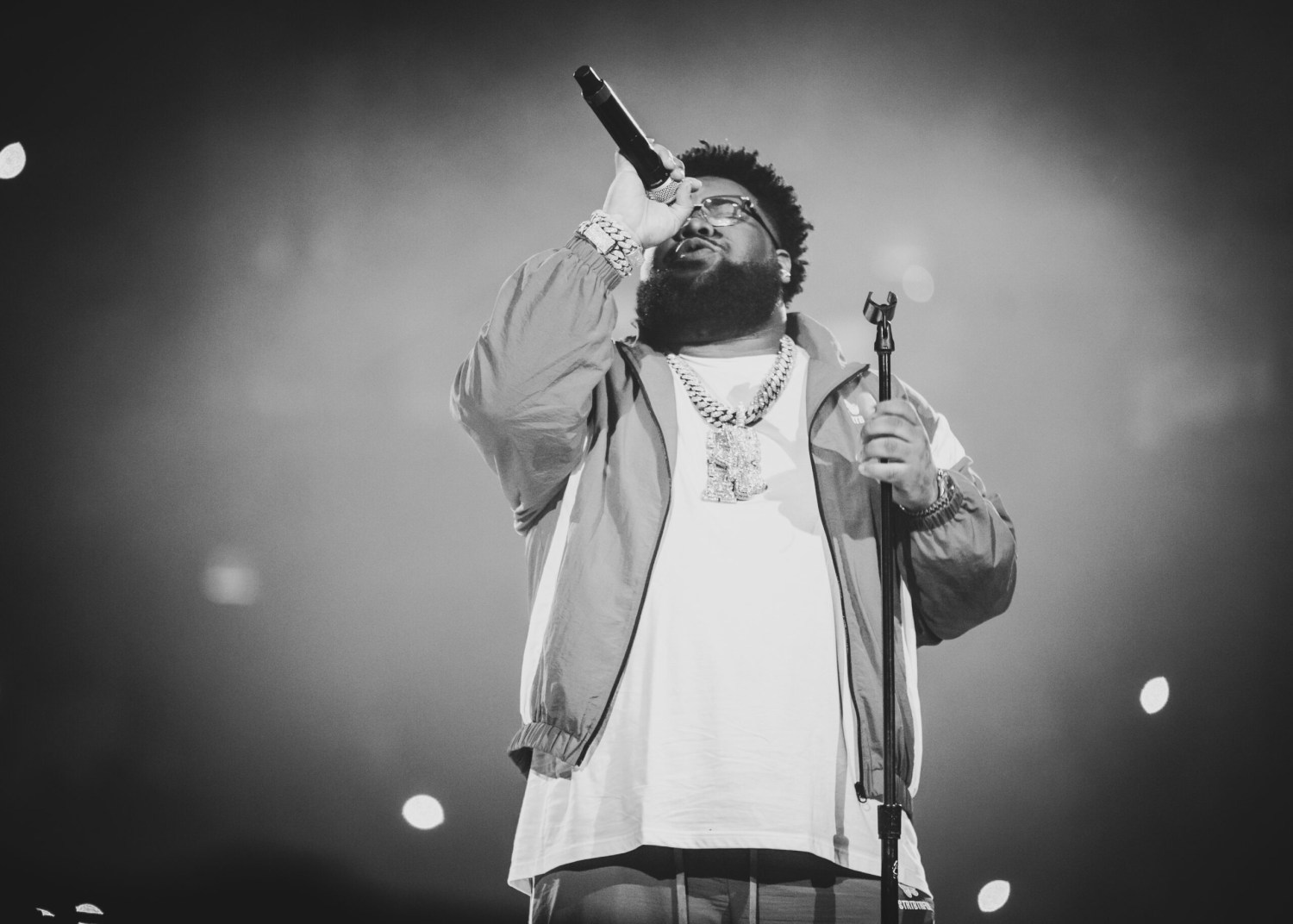 Man with beard and glasses holding a microphone up on stage, wearing chain necklaces and a jacket