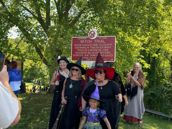 Three women and a child are dressed as witches and stand in front of a historical maker that says, "Legends & Lore, Witch Trial, Widow Krieger said to escape drowning in this river when tried as a witch ca. 1785. Accusers believed witches floated. She sank and was saved." They are smiling and posing for a photo.