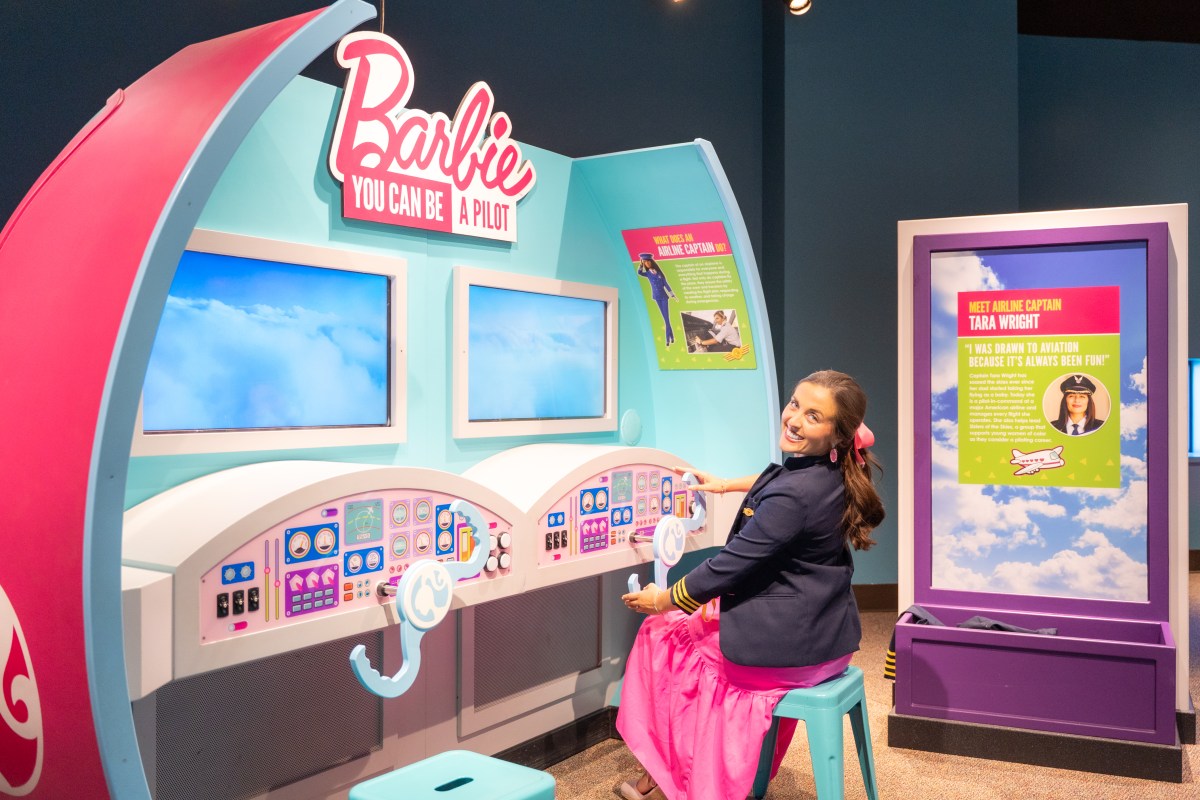 A woman sits at the Barbie You Can Be a Pilot cockpit display, exploring the exhibit featuring Captain Tara Wright