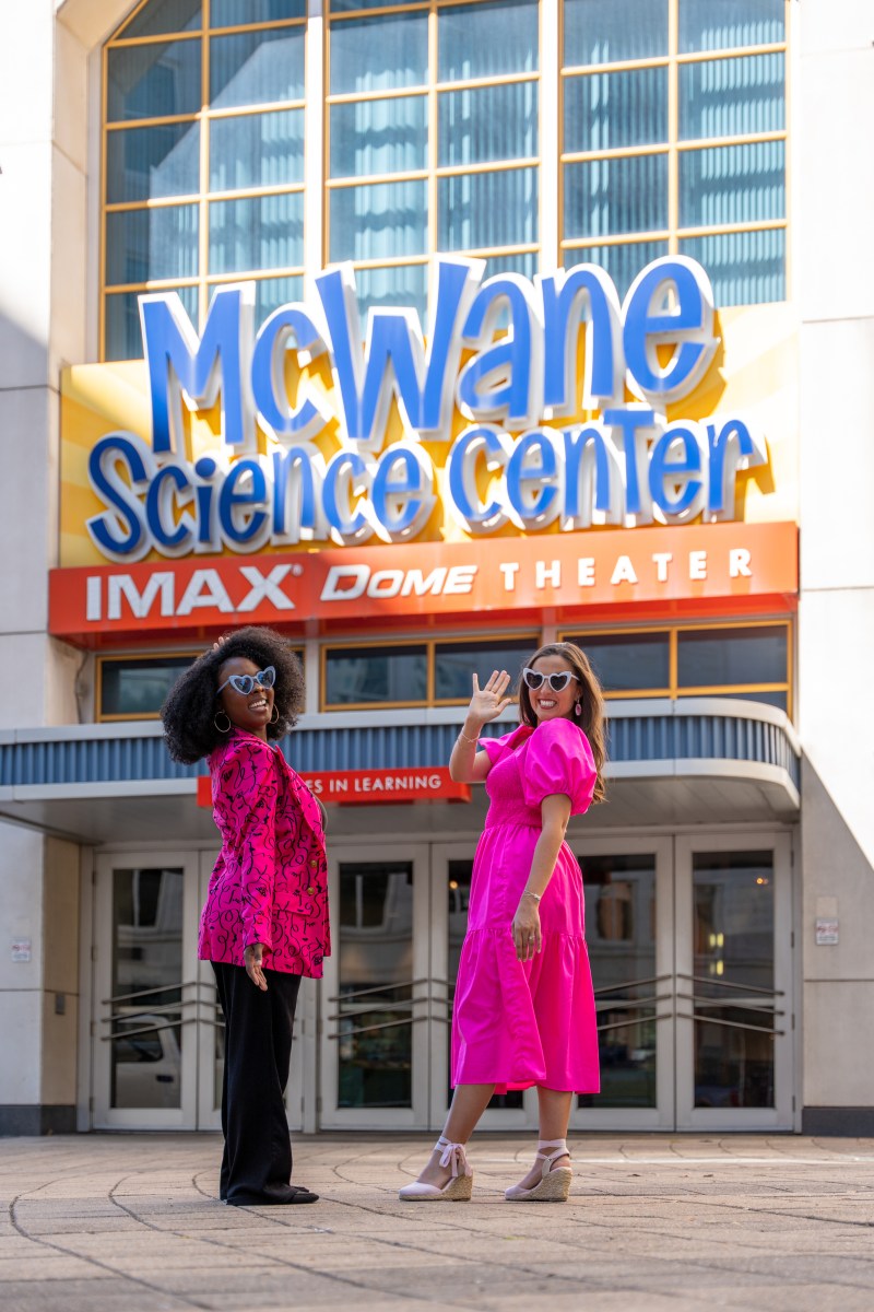 two young women stand smiling and waving in front of McWane Science Center in Birmingham