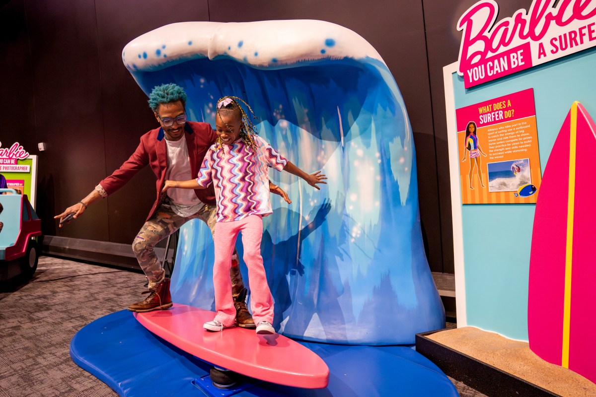 a father and daughter pretend to surf on a pretend surfboard on display at a "Barbie You Can Be A Surfer" exhibit