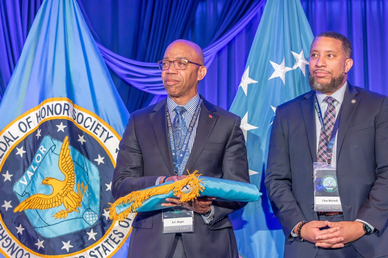 A.C. Roper holds a furled flag beside Chaz Mitchell at the 2026 Medal of Honor Convention, with the Medal of Honor Society seal displayed behind them