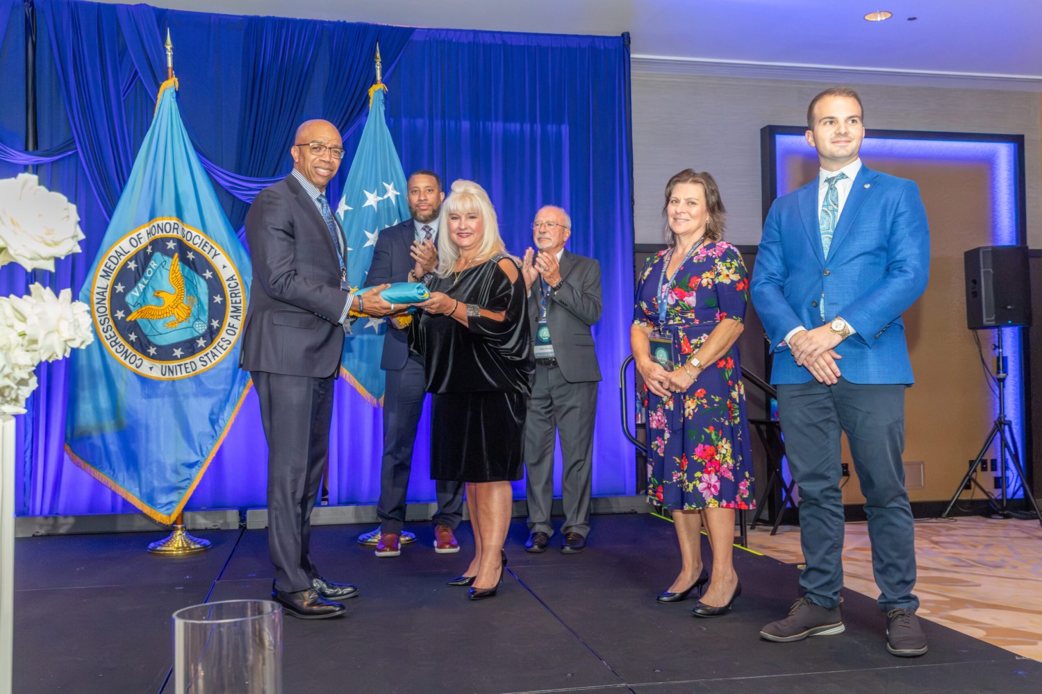 Man presents a wrapped gift to a woman on stage as others applaud, beside the Congressional Medal of Honor Society flag