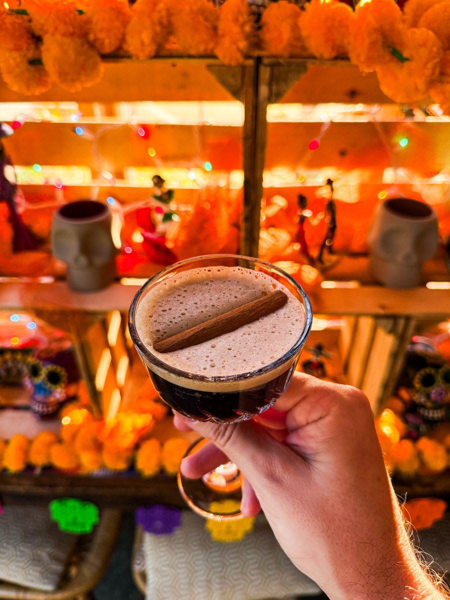 6 ways to celebrate Día de los Muertos in Birmingham 7 Wake the Dead cocktail with a cinnamon stick, held up in front of a marigold-decorated Día de los Muertos altar