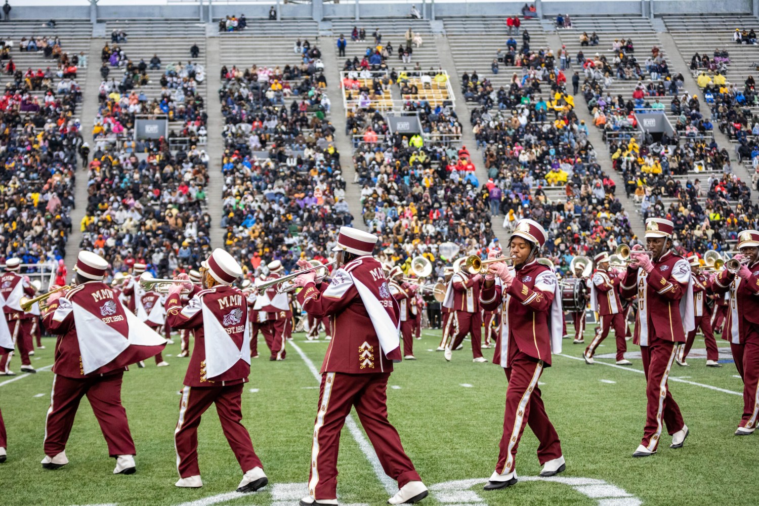 Marching band members in maroon and white uniforms play trumpets on a football field before a packed stadium crowd