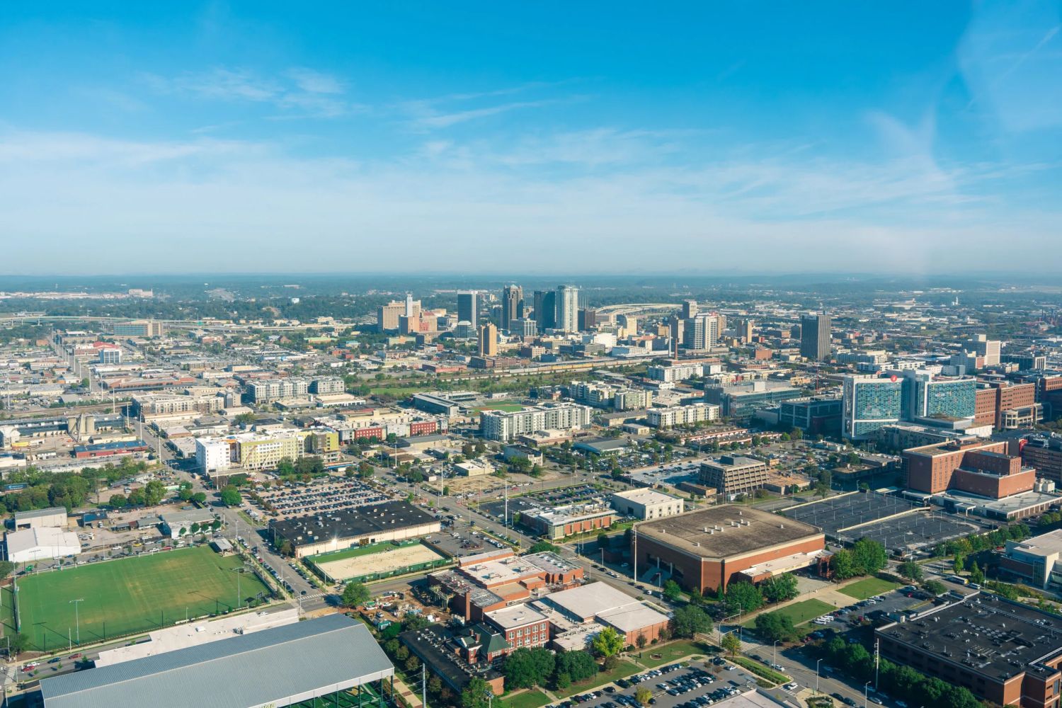 Aerial view of a city skyline with downtown skyscrapers surrounded by low-rise buildings, parking lots, and sports fields